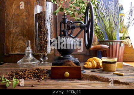 Rustikale Kaffeeszene mit Vintage-Mahlwerk, gerösteten Bohnen, gemahlenem Kaffee, Croissants und Grün auf einem Holztisch. Gemütliches Farmhouse-Frühstück im Stil Stockfoto