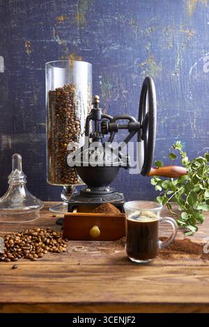 Eine Glasschale frisch gebrühten schwarzen Kaffees auf einem rustikalen Holztisch mit gemahlenem Kaffee, Bohnen und einem Vintage-Mahlwerk im Hintergrund, gemütlich gestaltet Stockfoto