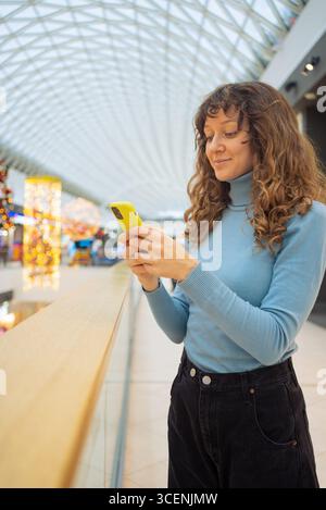 Lächelnde Frau, die ihr Handy benutzt, während sie im Einkaufszentrum steht, das für Feiertage dekoriert ist, genießt praktische digitale Kommunikation und festliche Atmosphäre Stockfoto