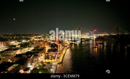 Blick aus der Vogelperspektive auf den beleuchteten River Street District mit seinen Backsteingebäuden und der Talmadge Memorial Bridge, die auf dem dunklen Fluss reflektiert, Savannah, Georgia, USA. Stockfoto