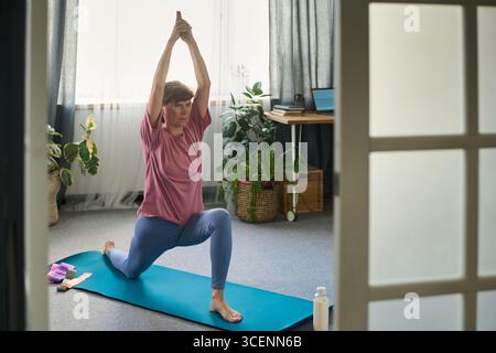 Ältere kaukasische Frau übt Yoga auf einer Trainingsmatte drinnen, mit erhobenen Armen, umgeben von Zimmerpflanzen und Fitnessaccessoires Stockfoto