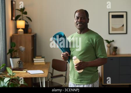 Senior Black man steht im Home Office mit Yogamatte und Wasserflasche, lächelnd und blickend nach vorne, Indoor-Wellness-Konzept mit Pflanzen und gerahmtem Kunstwerk im Hintergrund Stockfoto