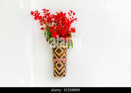 Bambusblumenvase mit roten Fliederblüten, die an der weißen Wand hängen. Isolierter Hintergrund. Stockfoto