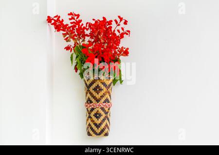 Bambusblumenvase mit roten Fliederblüten, die an der weißen Wand hängen. Isolierter Hintergrund. Stockfoto