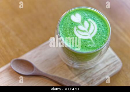 Matcha Latte mit herzförmiger Kunst aus Glas auf Holztablett. Heißer Matcha Latte mit zartem Herz und Blatt Latte Art serviert in doppelwandiger Glasschale Stockfoto