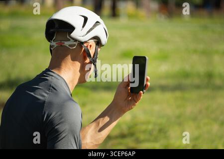 Männlicher Radfahrer, der ein Smartphone mit leerem Bildschirm auf dem Fahrrad im Park hält. Stockfoto