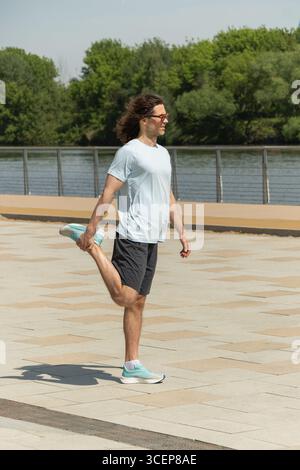 Gut aussehender Brünette Mann, der sportliche Freizeitkleidung und Sonnenbrille trägt, während er sich im modernen öffentlichen Park der Stadt warm-up-Übungen macht. Pause bei der Arbeit. Summe Stockfoto