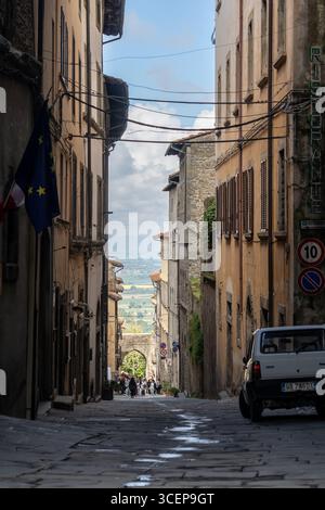 Cortona, Toskana, Italien - 24. April 2025: Blick auf eine enge Kopfsteinpflasterstraße, die von alten Gebäuden gesäumt ist, führt zu einem entfernten Bogengang und einem Blick auf die grüne toskanische Landschaft. Stockfoto
