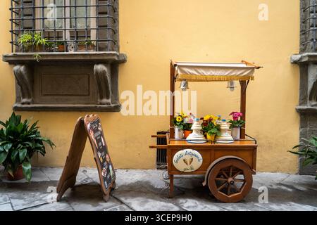 Tuscany, Italien - 24. April 2025: Blick auf einen bezaubernden Eiswagen mit leuchtenden Blumen, eingebettet an einer sonnendurchfluteten Mauer mit einem kunstvollen Fenster und einer rustikalen Menütafel. Stockfoto