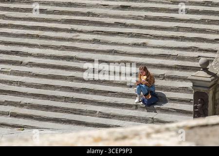 Cortona, Toskana, Italien - 24. April 2025: Blick auf eine Frau, die allein auf der Piazza Della Repubblica sitzt, in ihrem Buch inmitten der weitläufigen Steinstufen eingefangen und einen Moment der einsamen Betrachtung festgehalten. Stockfoto