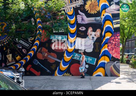 Brooklyn, USA - 13. August 2025: Blick auf ein lebendiges Wandbild mit einem Corgi, Musikinstrumenten und verschiedenen Objekten, während eine Frau vorbeigeht und eine dynamische Straßenszene schafft. Stockfoto