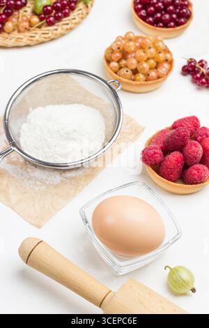 Mehl im Sieb, Ei in einer kleinen Glasschale. Tartlets mit Beeren. Weißer Hintergrund. Draufsicht Stockfoto