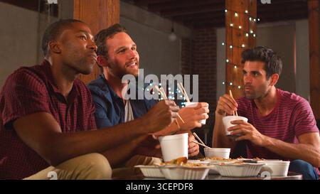 Eine Gruppe männlicher Freunde, die chinesische Mahlzeit zum Mitnehmen teilen und Fernsehen im Loft Apartment Stockfoto