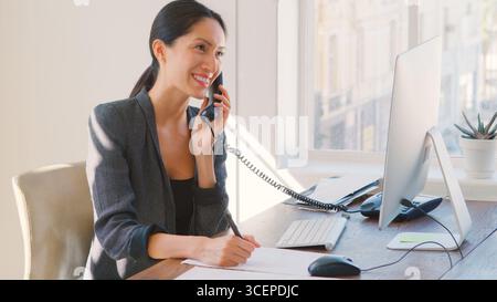 Junge Geschäftsfrau, Die Am Schreibtisch Am Computer Arbeitet Und Telefoniert Stockfoto
