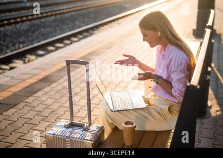 Elegante Geschäftsfrau mit Laptop und Ohrhörern, während sie am Bahnhof auf einer Bank mit Koffer sitzt. Sie spricht während eines Videoanrufs. Stockfoto