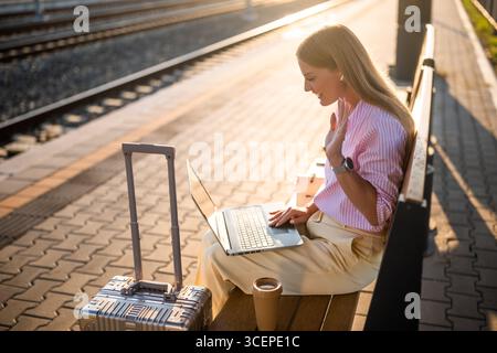 Elegante Geschäftsfrau, die Ohrhörer trägt und während eines Videogesprächs auf ihrem Laptop winkt, während sie mit Gepäck auf der Bank sitzt und auf die Ankunft des Zuges wartet Stockfoto