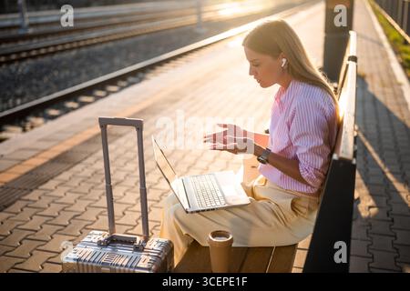 Elegante Geschäftsfrau mit Laptop und Ohrhörern, während sie am Bahnhof auf einer Bank mit Koffer sitzt. Sie spricht während eines Videoanrufs. Stockfoto