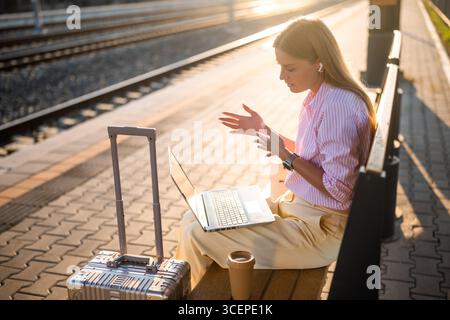 Elegante Geschäftsfrau mit Laptop und Ohrhörern, während sie am Bahnhof auf einer Bank mit Koffer sitzt. Sie spricht während eines Videoanrufs. Stockfoto