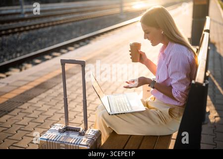Geschäftsfrau, die auf einer Bank am Bahnhof mit Koffer sitzt, Laptop und Kopfhörer benutzt, während sie Kaffee trinkt. Sie hat einen Videoanruf. Stockfoto