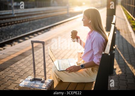 Geschäftsfrau, die auf einer Bank am Bahnhof mit Koffer sitzt, Laptop und Kopfhörer benutzt, während sie Kaffee trinkt. Sie hat einen Videoanruf. Stockfoto
