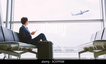 Junger Geschäftsmann, Der In Der Airport Departure Lounge Sitzt Und Das Handy Benutzt, Um Den Flugzeugstart Zu Beobachten Stockfoto