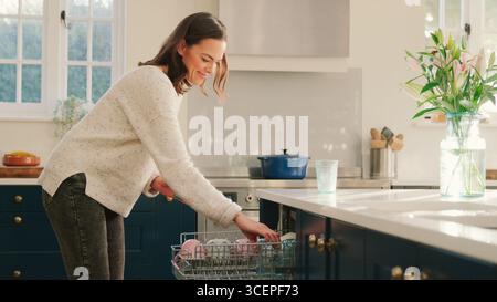 Frau Zu Hause In Der Küche, Die Hausaufgaben Macht Und Gläser In Den Geschirrspüler Lädt Stockfoto