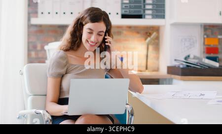 Junge Lächelnde Geschäftsfrau, Die Am Schreibtisch Im Büro Arbeitet Und Mit Dem Handy Spricht Stockfoto