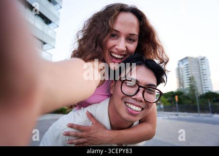 Selfie eines jungen Paares, das in die Kamera schaut, während der Freund seiner Freundin Huckepack gibt. Stockfoto