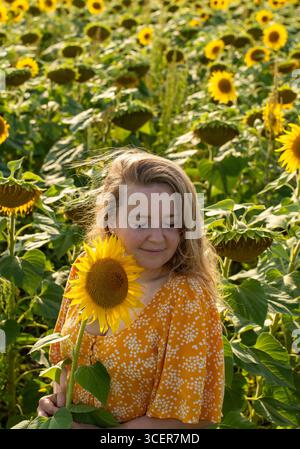 Schöne blonde Frau im orangen Blumenkleid, die Sonnenblume mit geschlossenen Augen in blühendem Sonnenblumenfeld am sonnigen Sommertag hält. Naturbegriff Stockfoto