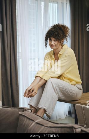 Niedliche Frau mit lockigen Haaren sitzt lässig auf einem Tisch in einem gut beleuchteten Raum. Sie trägt ein gelbes Hemd und eine graue, lose Hose, die eine entspannte Atmosphäre vermittelt Stockfoto