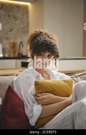 Eine junge Frau mit lockigen Haaren sitzt bequem auf einer Couch in einem stilvollen Wohnzimmer. Sie umarmt ein gelbes Kissen und trägt ein lässiges Outfit, das als Rela erscheint Stockfoto
