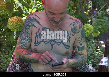 Junger Mann auf Handy, mit Tätowierungen auf Armen und Oberkörper, sitzt unter einem roten Sonnenschirm im Garten, Baden-Württemberg, Deutschland Stockfoto