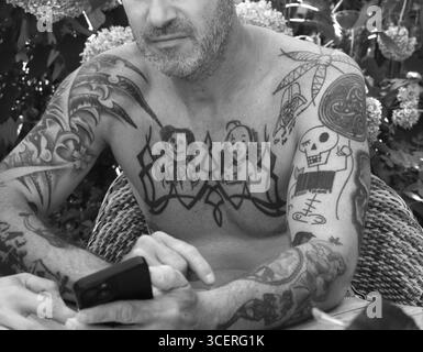 Junger Mann auf Handy und Tätowierungen auf Armen und Oberkörper, im Garten sitzend, schwarz-weiß, Baden-Württemberg, Deutschland Stockfoto