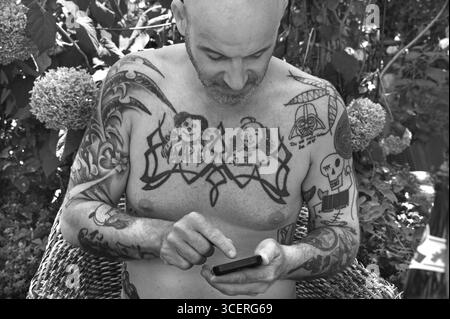 Junger Mann auf Handy, mit Tätowierungen auf Armen und Oberkörper, sitzt unter einem Sonnenschirm im Garten, schwarz-weiß, Baden-Württemberg, Deutschland Stockfoto