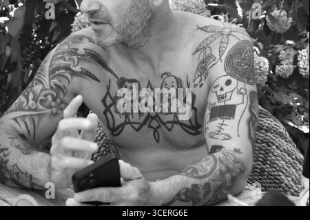 Junger Mann auf Handy und Tätowierungen auf Armen und Oberkörper, im Garten sitzend, schwarz-weiß, Baden-Württemberg, Deutschland Stockfoto