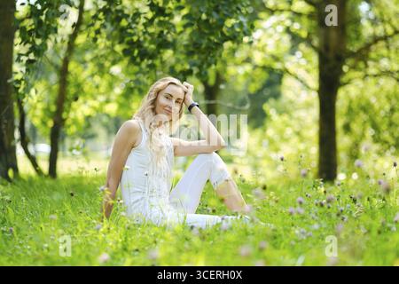 Eine Frau mit langen blonden Haaren sitzt auf weichem Gras, umgeben von blühenden Blumen in einem lebhaften Park. Das Sonnenlicht zieht durch Bäume und schafft ein ruhiges A Stockfoto
