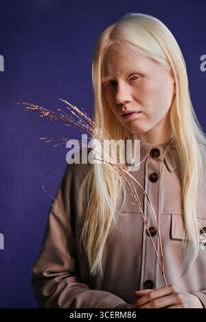 Vertikaler Schuss einer schönen jungen kaukasischen Frau mit Albinismus, die ein lässiges Hemd trägt, das eine vergoldete Pflanze hält, die vor violettem Hintergrund steht Stockfoto