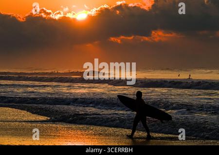 Isle Of Palms, Usa. Januar 2016. Ein Surfer im Sonnenaufgang spaziert entlang der Wasserlinie auf der Suche nach der perfekten Welle am Front Beach, 19. August 2025 in Isle of Palms, South Carolina. Hohe Wellen und gefährliche Fluten treffen die Region, als Hurrikan Erin, ein Sturm der Kategorie 2, vor der Küste vorbeizieht. Quelle: Richard Ellis/Richard Ellis/Alamy Live News Stockfoto