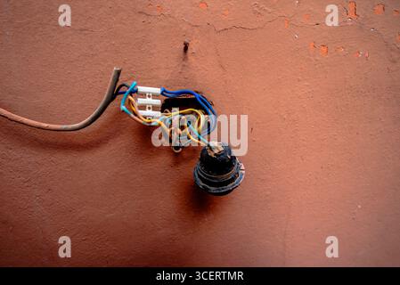Nahaufnahme eines freiliegenden elektrischen Lampenhalters mit verwickelten Drähten an einer abgenutzten Terrakotta-Wand. Ein düsteres städtisches Detail, das die alternde Infrastruktur und widerspiegelt Stockfoto