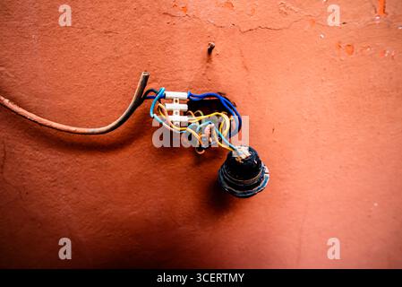 Freiliegender Lampenhalter mit verwickelten farbigen Drähten an einer strukturierten Terrakotta-Wand. Eine Nahaufnahme der veralteten Infrastruktur, Straßenreparatur und urbanen Util Stockfoto