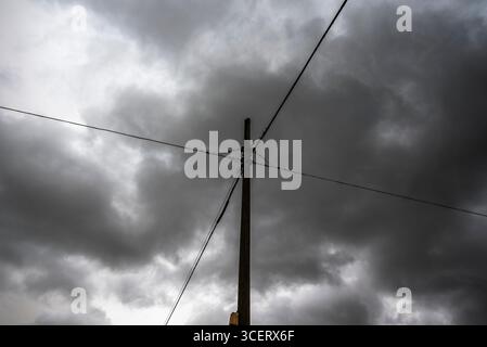 Silhouette eines Versorgungsmasts mit sich kreuzenden Kabeln, die ein Kreuzmuster gegen einen stimmungsvollen, bewölkten Himmel bilden. Ein dramatisches Bild, das Spannung symbolisiert, Conne Stockfoto
