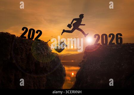 2026, Willkommen Frohe Weihnachten und Happy New Year 2026 Silhouette Mann springt von den Klippen von 2025 zu den Klippen von 2026 mit dem Himmel und den Strahlen von t Stockfoto