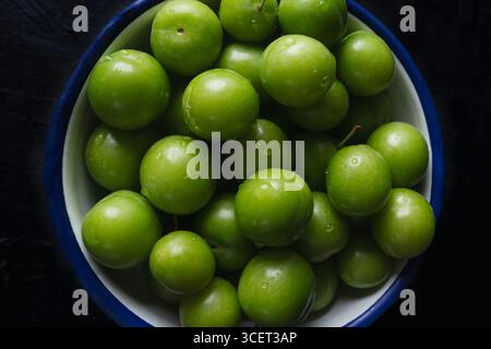 Nahaufnahme von frischen grünen Pflaumen in einer weißen Schüssel mit blauem Rand auf dunklem Hintergrund. Stockfoto