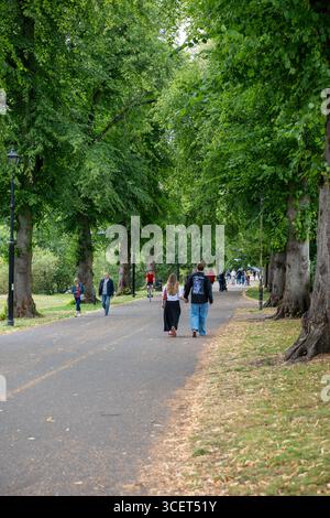 Ein Paar geht Hand in Hand auf einem von Bäumen gesäumten Weg in einem Park, umgeben von üppigem Grün und anderen Menschen, die den Tag genießen. Stockfoto
