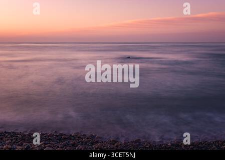Ein einsamer Sonnenaufgang entfaltet sich an einem friedlichen Strand mit kleinen Steinen, mit glattem Wassereffekt und warmen Orangetönen, die einen Sinn für ein Gefühl erwecken Stockfoto