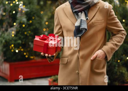 Herren in beigefarbenem Mantel, die eine rote Weihnachtsschachtel in der Nähe des Weihnachtsbaums auf dem Stadtmarkt halten. Weihnachtseinkäufe und frohe Feiertage. Im Freien. Nahaufnahme. Stockfoto