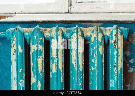 Nahaufnahme eines alten Gusseisenheizkörpers mit abblätternder türkisfarbener und gelber Farbe, der an einem verwitterten Wand- und Fensterrahmen angebracht ist. Das rustikale Textu Stockfoto