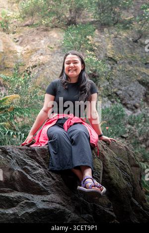 Indonesische Frau sitzt auf einem Felsen mit einem großen tropischen Wasserfall im Hintergrund. Stockfoto