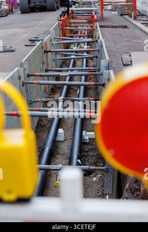 Städtischer Straßenaushub mit sichtbaren Rohrleitungen und Sicherheitsbarrieren. Rote Warnleuchten und Bauzäune markieren den Standort in einem Wohngebiet Stockfoto