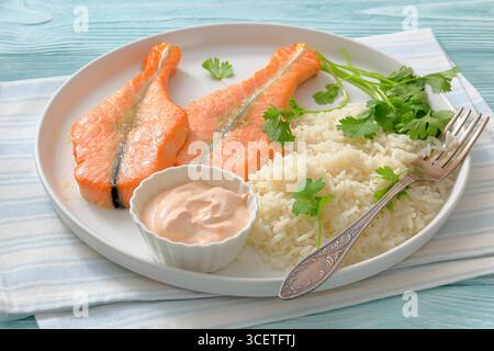 Gebackene Lachssteaks mit Reis, Koriander und cremiger Sauce auf einem weißen Teller auf Holztisch mit Gabel und Serviette, Landschaftsblick von oben, nicht AI Gattungen Stockfoto
